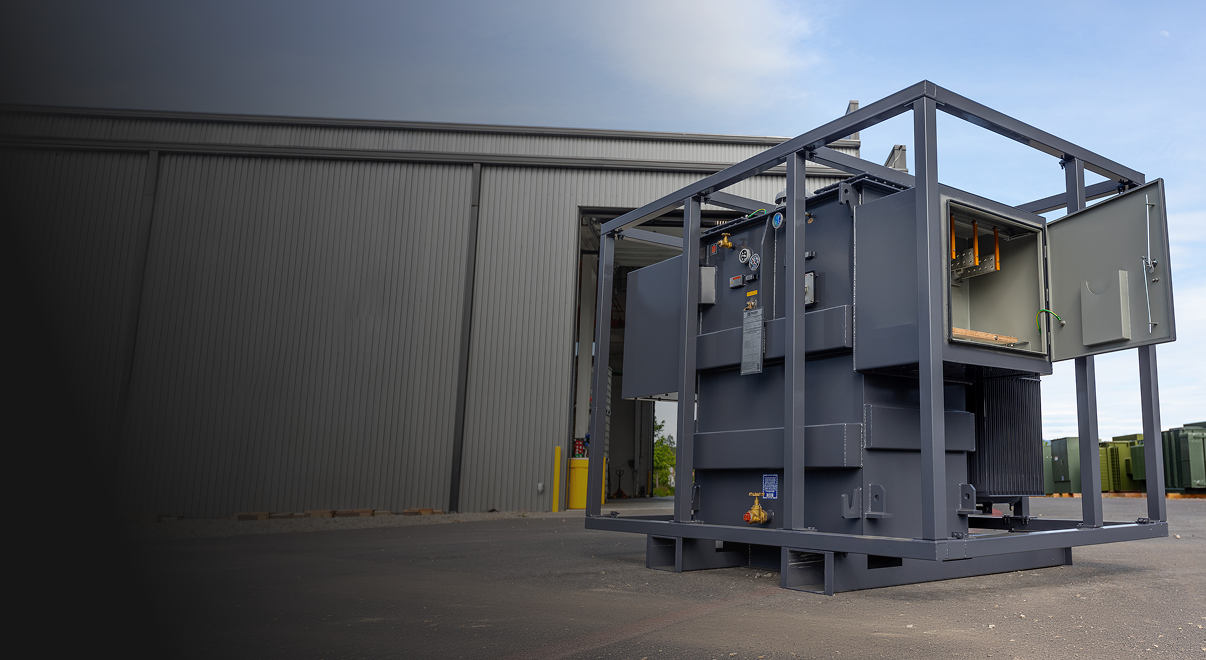 Large gray industrial rental transformer outdoors near a warehouse with an open panel door.